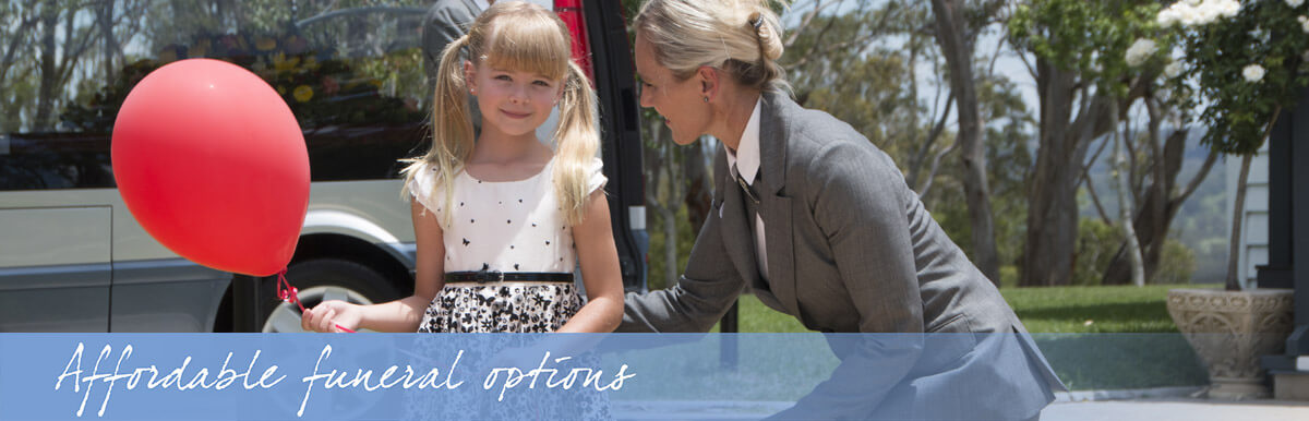 A funeral director kneeling to speak gently with a young girl holding a red balloon