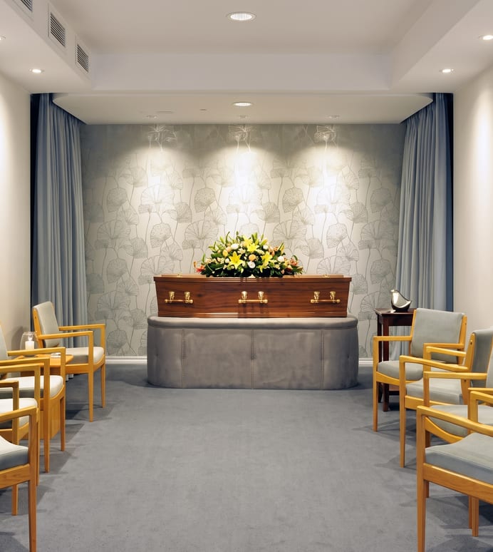 A softly lit funeral viewing room with a flower-topped coffin on a padded platform, patterned wallpaper behind it, and timber chairs arranged in rows facing it