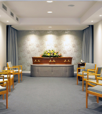 A softly lit viewing room with a flower-topped coffin on a catafalque, timber chairs arranged around it