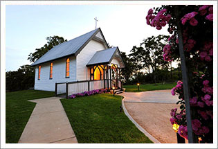 A small white timber chapel lit at dusk, surrounded by rose bushes and a winding garden path