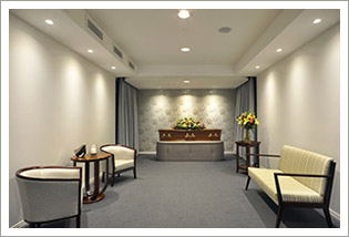 A softly lit viewing room with a flower-topped coffin on a catafalque, chairs arranged around it